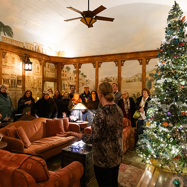 The owner of the mission inn is leading a christmas candlelight house tour in her living room with a beautiful Christmas tree lit up behind her