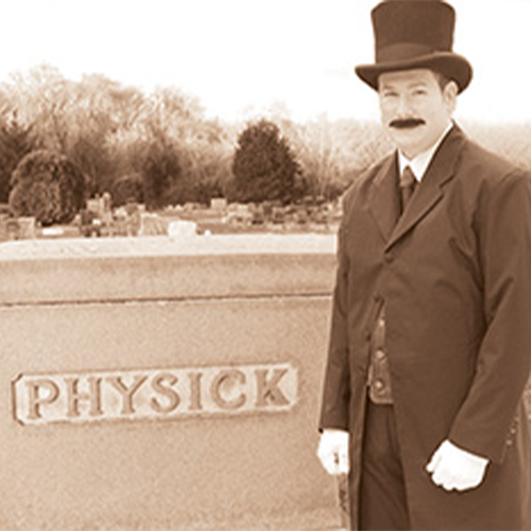 Doctor Emlen Physick dessed in a top hat and victorian coat stnading next to his gravestone that says Physick