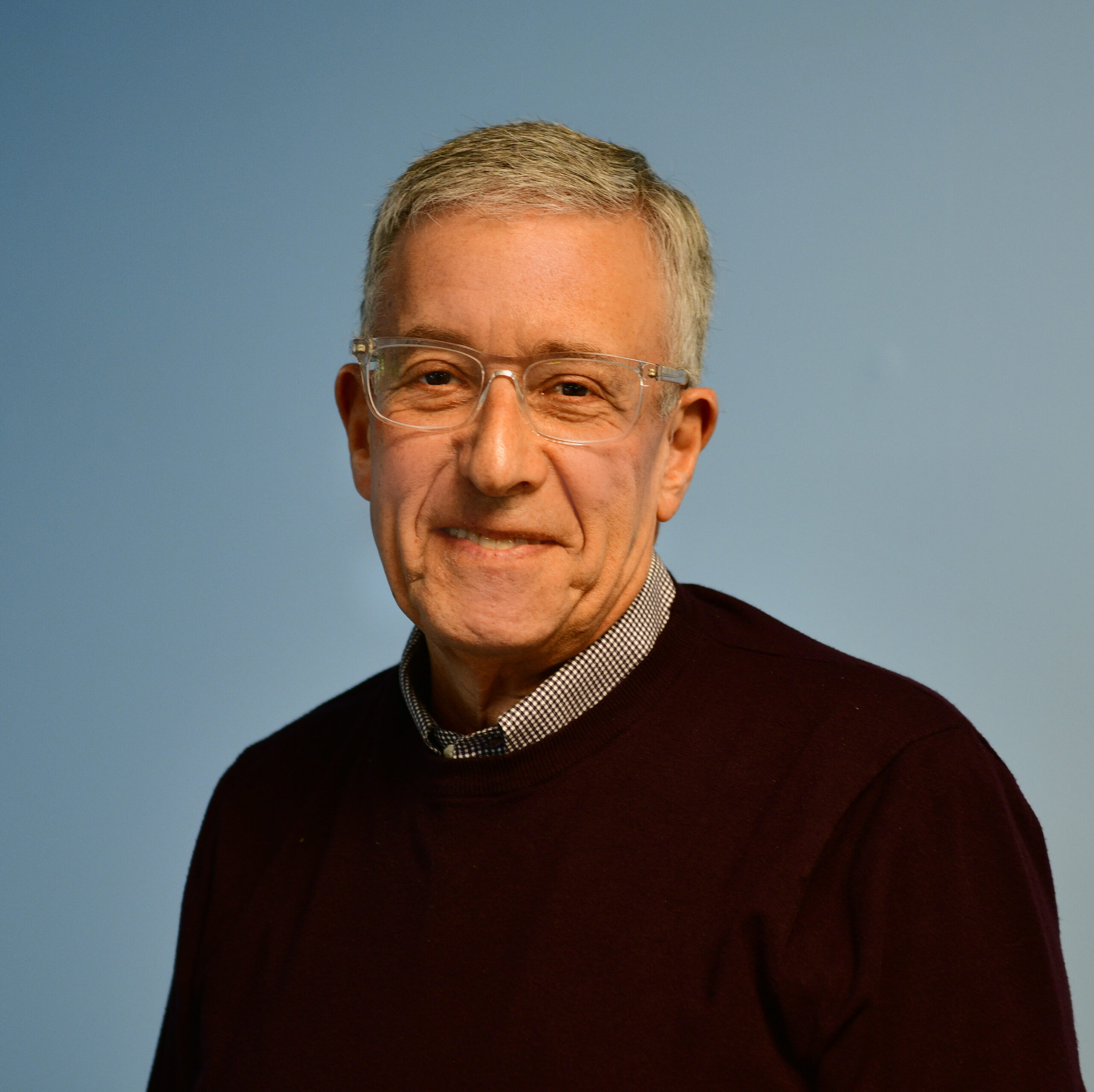 a headshot of a white man in glasses in front of a blue backdrop